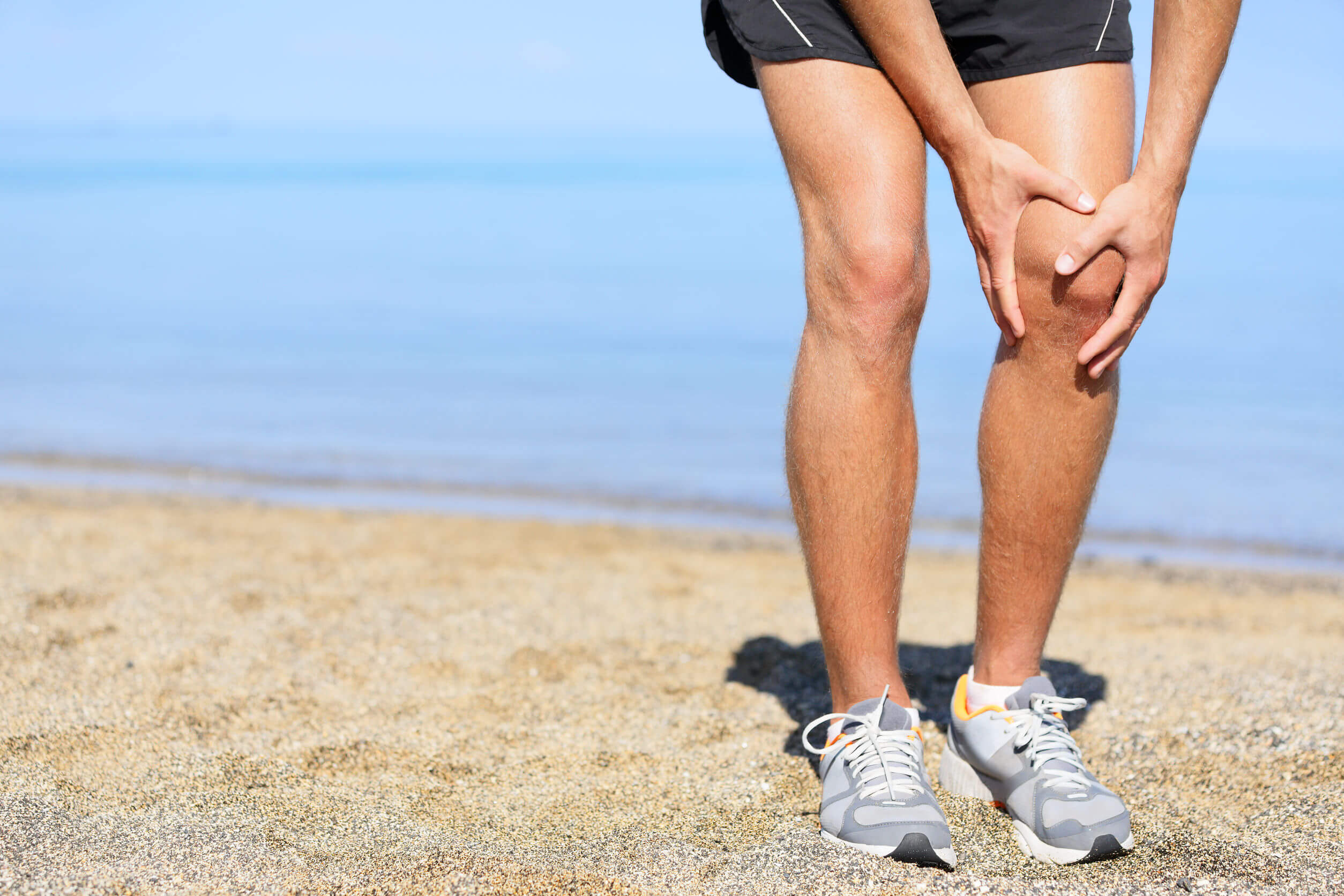 running injury - man jogging with knee pain. close-up view of runner injured jogging on the beach clutching his knee in pain. male fitness athlete.