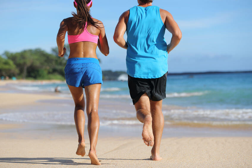 Two athletes runners couple running together on beach. People from behind jogging away barefoot on sand on tropical travel destination. Lower body, legs, feet.