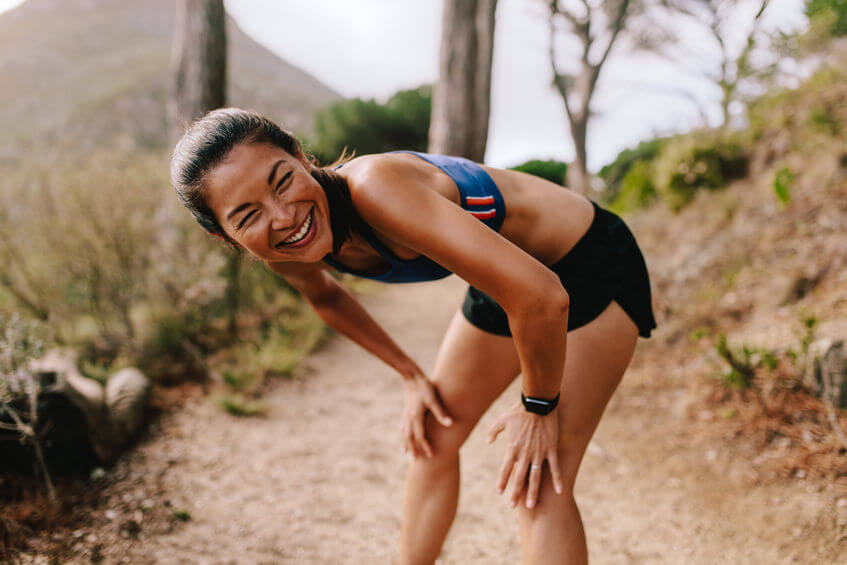 Fit young asian woman standing on mountain trail with her hands on knees and smiling. Female runner in sportswear taking a break after running workout.